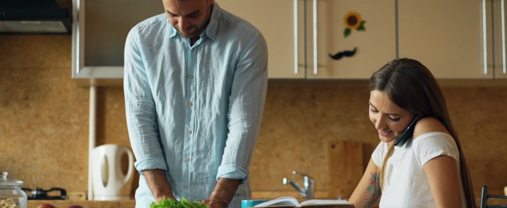 Attractive couple in the kitchen early morning. Beautiful girl chatting phone while her boyfriend cooking breakfast at home